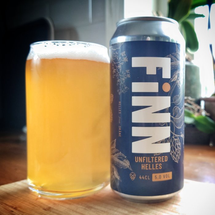 Ett glas med ljust öl bredvid en ölburk med texten "FINN UNFILTERED HELLES" på en träyta.