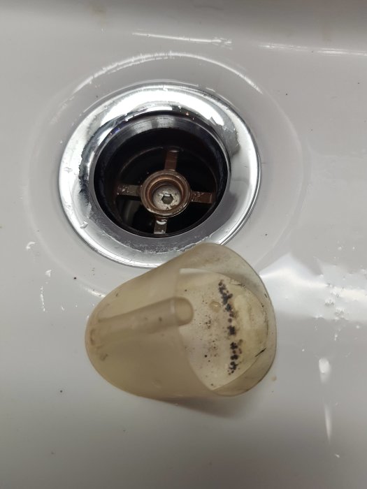 En smutsig vit silikonkil bredvid ett öppet avlopp i ett handfat, med fokus på den borttagna delen.