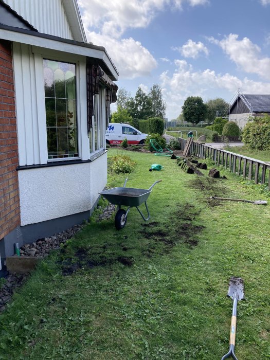 Trädgård med utsikt mot gatan, grävd mark för fiberinstallation och en tom skottkärra, bredvid ett hus.