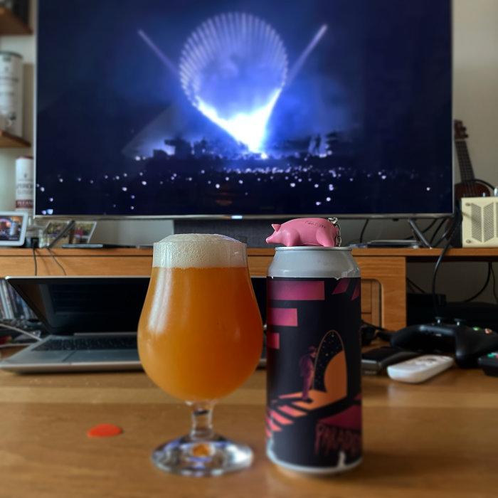 Ölglas och burk med Pink Floyd-tema framför TV som visar konsert, liten plastgris ovanpå burken.