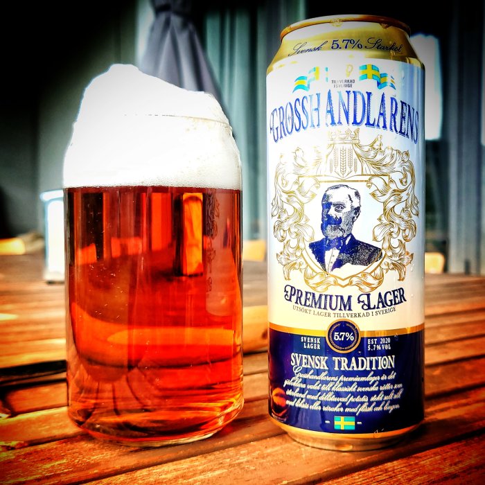 Ölglas med rikligt skum bredvid en burk av "Grosshandlarens Premium Lager" på ett träbord.