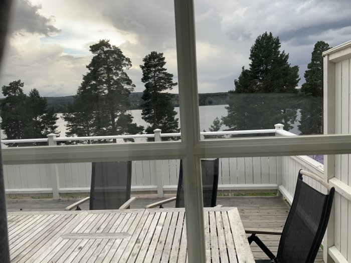 Utsikt från en veranda med trägolv och vit räcke över en sjö med skogsbeklädda öar och molnig himmel.