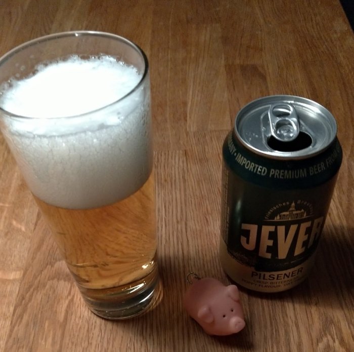 Jever ölburk bredvid ett glas fullt av skummande öl och en liten plastgris på ett träbord.