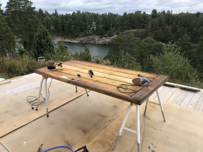 Träbord av teak under bearbetning utomhus med verktyg, mot skog och sjö.