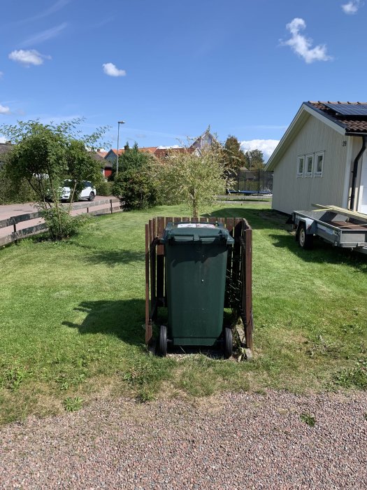 Nytt träskydd för soptunna i en trädgård framför villor under en klar himmel.