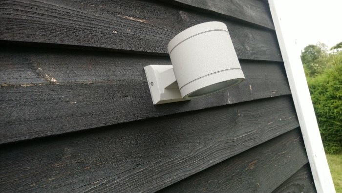 Cylindrisk fasadarmatur monterad på mörk trävägg med G9-sockel inuti.
