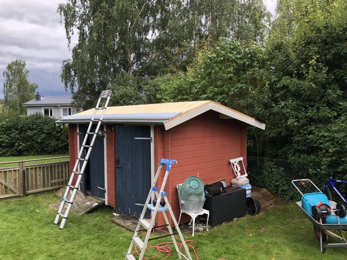 Röd uthusbyggnad med ny hängränna och otäckt tak, stege lutad mot byggnaden, trädgårdsredskap i förgrunden.