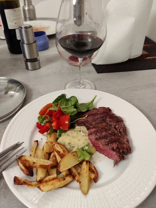 Tallrik med entrecote, klyftpotatis, sallad och sås, ett glas rödvin i bakgrunden.