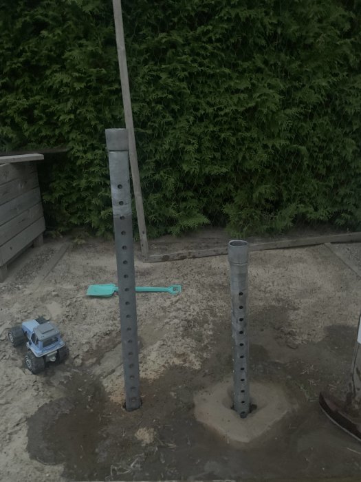 Två metallstolpar i marken med hål, ena utan spets, i en trädgård med leksakstruck och verktyg.