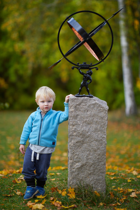 En liten pojke står bredvid en grå stenstolpe med en skulptur ovanpå i en park med höstlöv på marken.