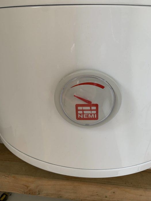 Termometer på en varmvattenberedare med röd visare och logotyp, fråga om dess funktion.