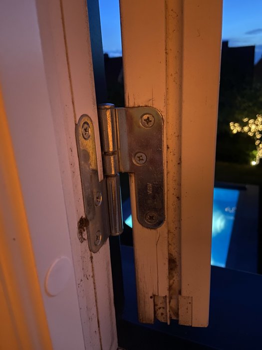 Närbild på ett fönsterbeslag mellan en öppen fönsterbåge och karm vid skymningen, med en pool i bakgrunden.