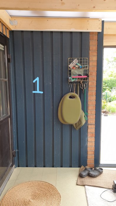 Del av ett uterum med isolerat golv och mörkblått panelat vägg segment 1 intill tegelstolpe, med redskapshängare och skor.