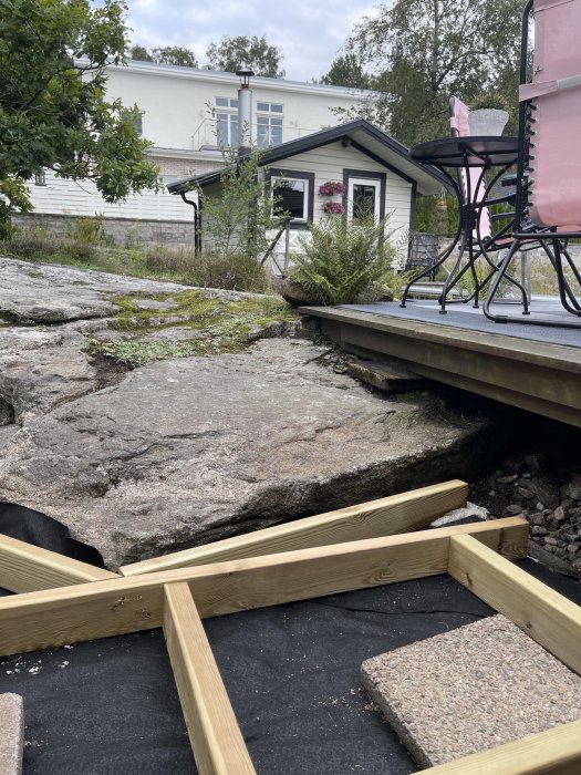 Uteplats under konstruktion med träramar och stenplattor framför ett vitt hus och en trädgård.