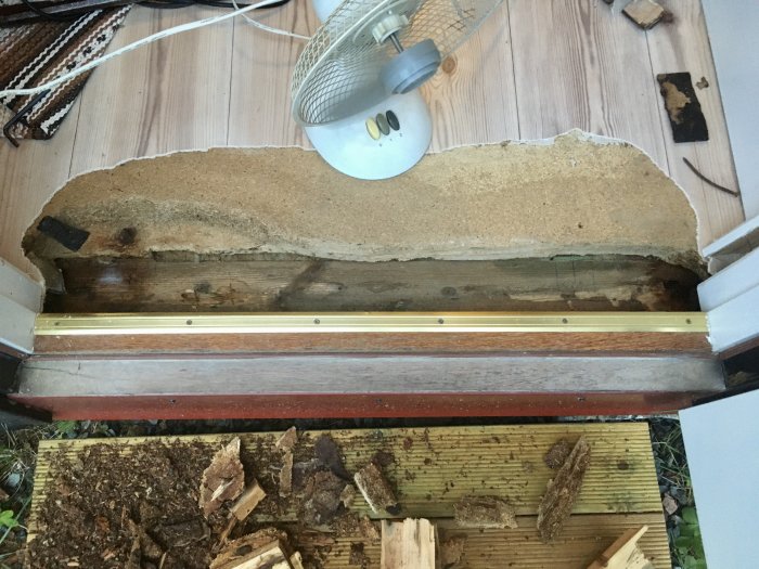 Uppskuret golvspån vid en ytterdörr som avslöjar en underliggande träbjälke med tecken på vattenskada och myrbo.