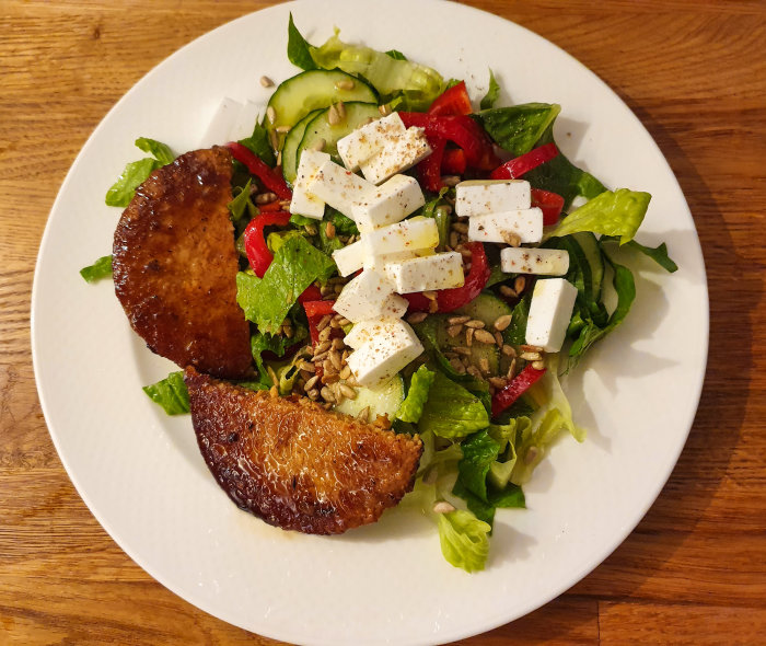 En tallrik med sallad, fetaost, gurka, röd paprika, solrosfrön och två grillade kycklinglår.