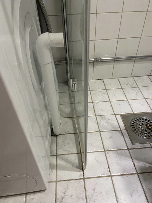 Badrum med vit tvättmaskin ansluten till synligt avloppsrör vid duschen, med tanke på möjlig optimering.