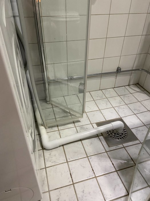 Snett perspektiv på ett badrumsgolv med vit klinker, där en synlig PVC-avloppsrör leder till ett golvbrunn nära duschhörna.
