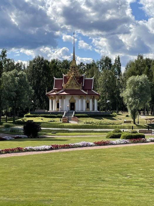 Thailändskt tempel med gyllene spira i Bispgården, omgivet av gröna gräsmattor och blomsterarrangemang.