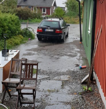 Vattenpölar nära husgrund med en bil parkerad, vattenflöde synligt på en grusig uppfart.