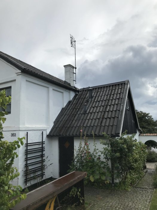 Vitputsat hus med svart tak och skorsten, omgivet av gröna buskar och en grå himmel.