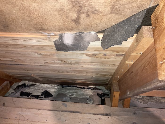 Ljusa fläckar på takpapp och råspont i vind med synliga träbjälkar och isoleringsmaterial.