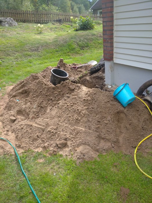 Uppgrävd hörna av en trädgård med synliga rör och sand, nära husvägg och gräsmatta.