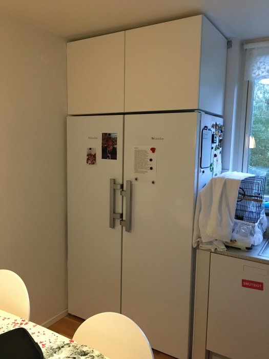 Kök med Mieles kyl och frys samt Ikea överskåp monterade ovanför, dekorerade med magneter och anteckningar.