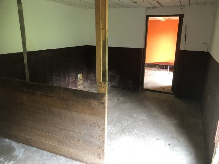 Interiör av ett gammalt stall med betonggolv och väggar, träpartition, öppen dörr till ett annat rum.