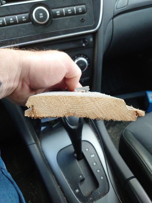 Hand håller en trasig bit träpanel i bilen, söker information om paneltypen för utbyte.