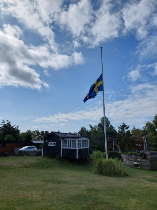 Svensk flagga vajar på en flaggstång framför en typisk trästuga med glasveranda i en trädgård.