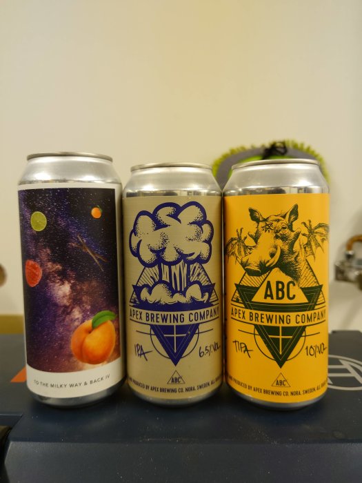 Tre olika ölburkar från Apex Brewing Company med konstnärliga etiketter på en bordsskiva.