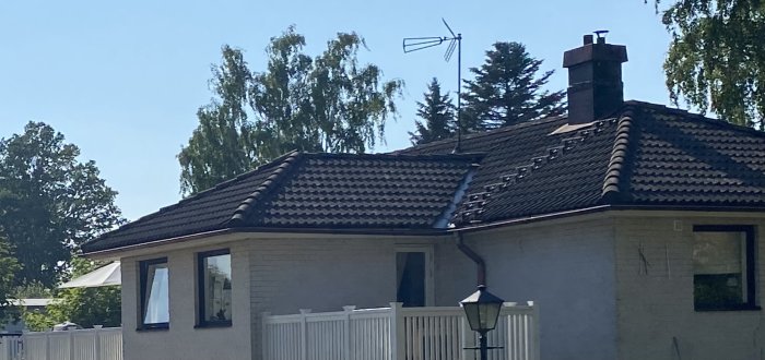 Ett hus med valmat tak och en antenn på taket, omgivet av träd och en blå himmel.