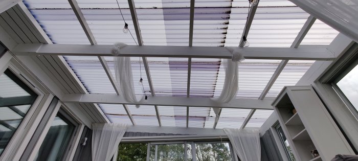 Utsikt underifrån av ett tak med genomskinliga takplattor och vita takbjälkar i en veranda.