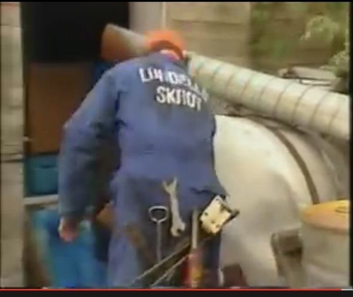 Person i blå arbetsklädsel och hjälm bredvid stor rörledning med texten "Lim, Olja, Skit" på ryggen.