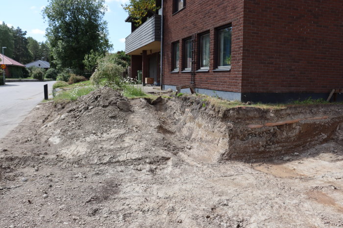 Grävd mark vid hus med synlig grund och balk längs asfalterad väg.