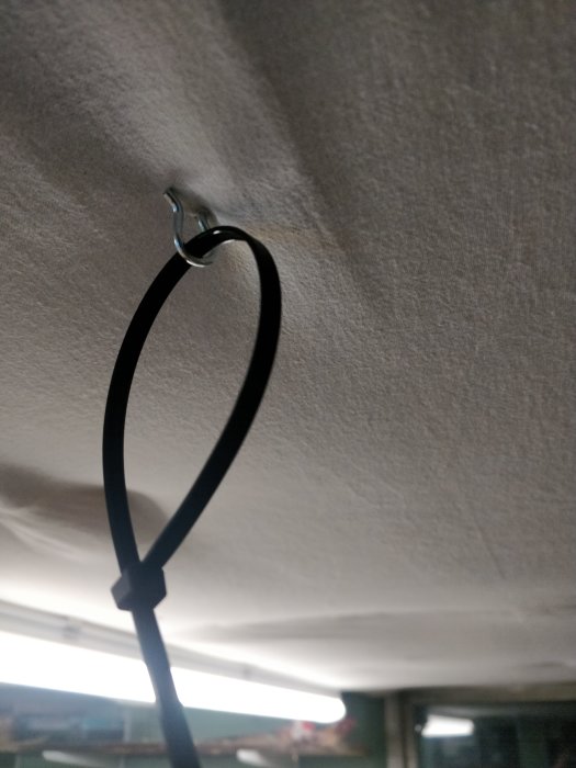 En krok i taket med en svart rem fäst i den, som håller ett vitaktigt skydd upphängt under arbetsytan.