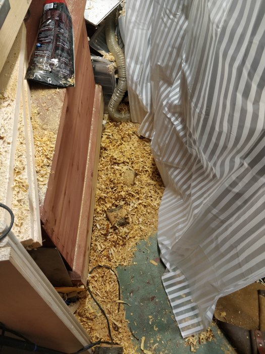 Stökigt arbetsutrymme med träspån på golvet och ett randigt duschdraperi hängandes i verkstaden.