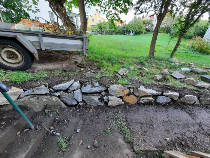 Nybyggd mur av sprängsten i en trädgård med en släpkärra och grönt gräs i bakgrunden.