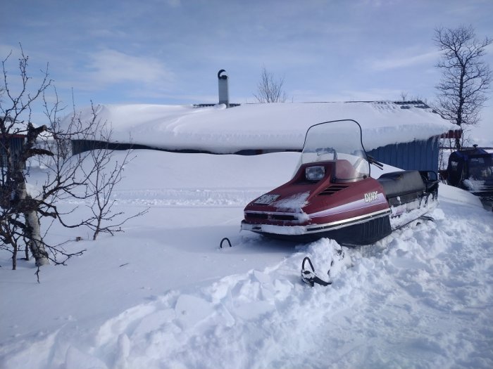 En snöskoter parkerad i djup snö med ett snötäckt hus i bakgrunden.