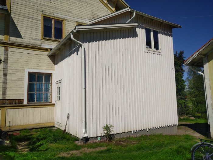 Nyrenoverat utbyggnad med vit panel, oputsad grunddel, och takavvattningssystem under en klarblå himmel.