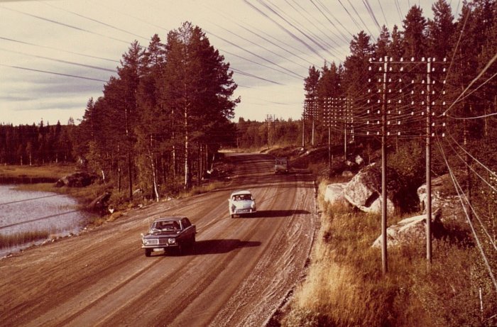 Gamla telefonstolpar och trådar längs en grusväg med två bilar från 1900-talet, omgivna av natur.