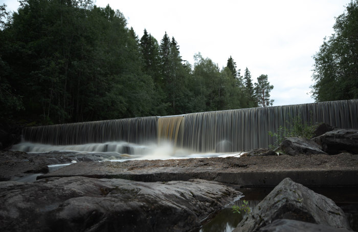 Ett konstgjort vattenfall med mjukt flödande vatten, omgivet av gröna träd och stenar i förgrunden.