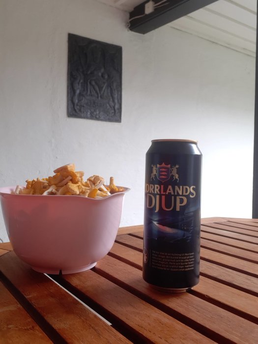 En skål med svamp och en burk Norrlands Djup öl på ett träbord utomhus.
