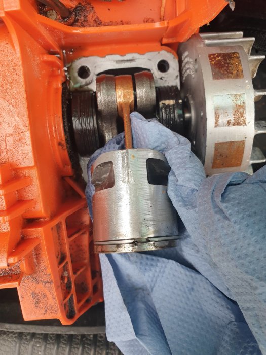 Hand i skyddshandske håller en motorcylinder mot bakgrund av orange maskindelar.