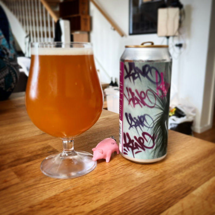 Ett glas öl bredvid en graffitidekorerad ölburk och en liten rosa grisfigur på ett träbord.