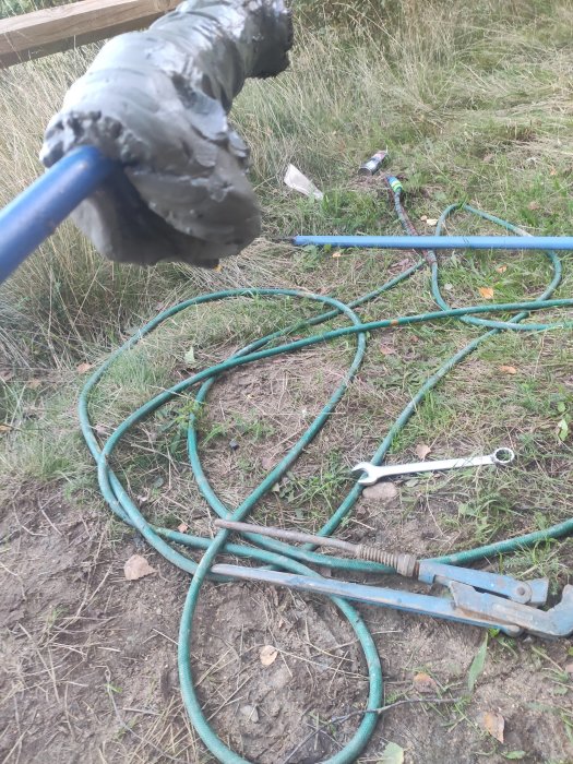 Styv lera på en borr som hålls upp, med trädgårdsutrustning och en slingrig vattenslang på marken i bakgrunden.