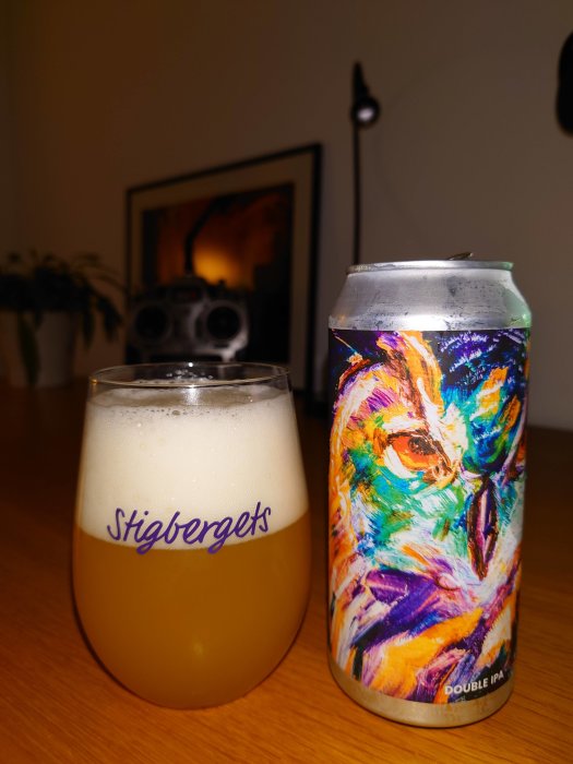 Ölglas med skum och färgrik Double IPA burk på bord framför suddig stereo och inramad bild.