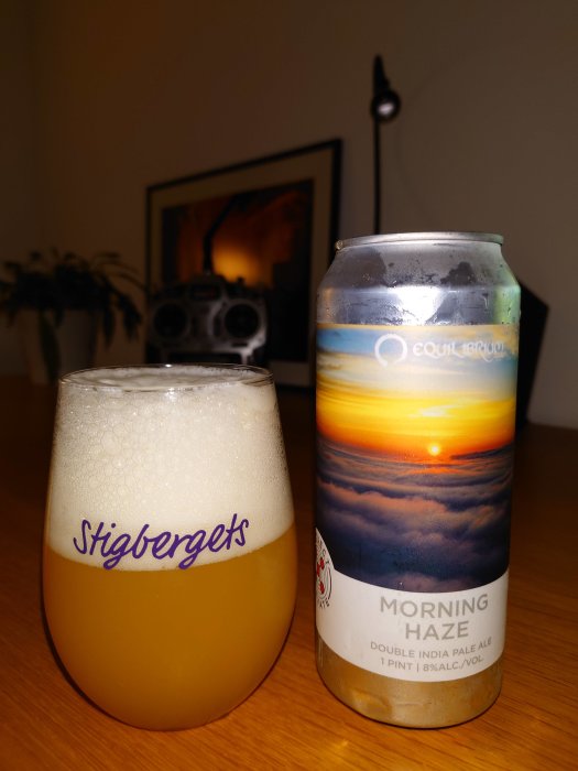 Ett glas öl och en burk med etiketten 'Morning Haze' på ett bord, med suddig bakgrund.