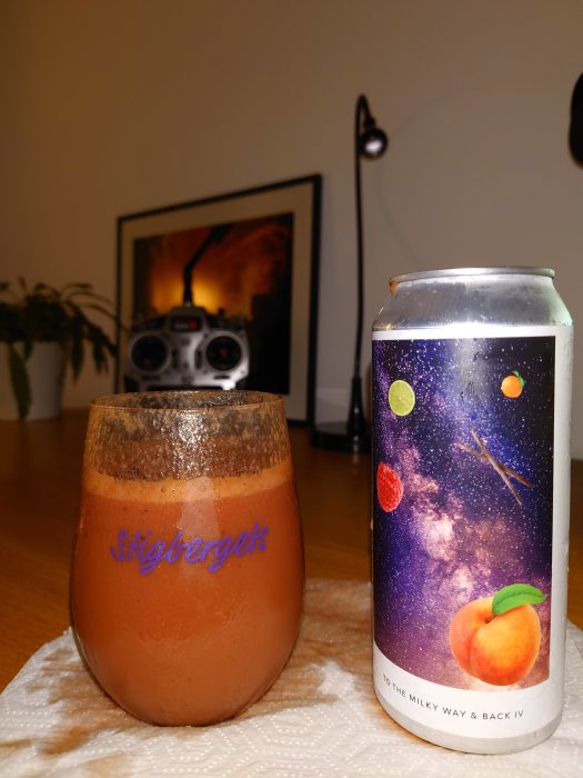 Ett ölglas med texten "Stigbergets" fyllt med en mörk dryck, bredvid en ölburk med rymdmönster och en persika. Bakgrund med radio och tavla.